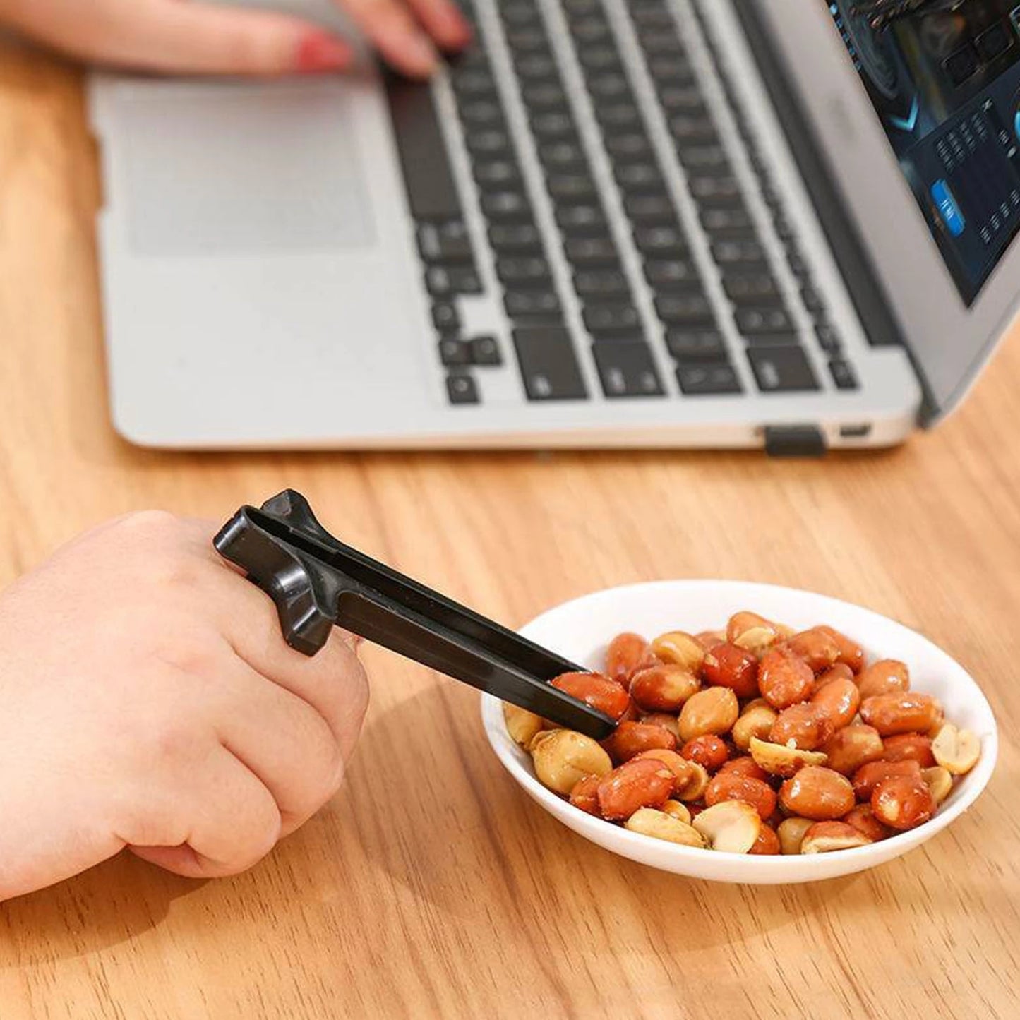 Person using tongs to pick up peanuts from a bowl on a desk with a laptop in the background