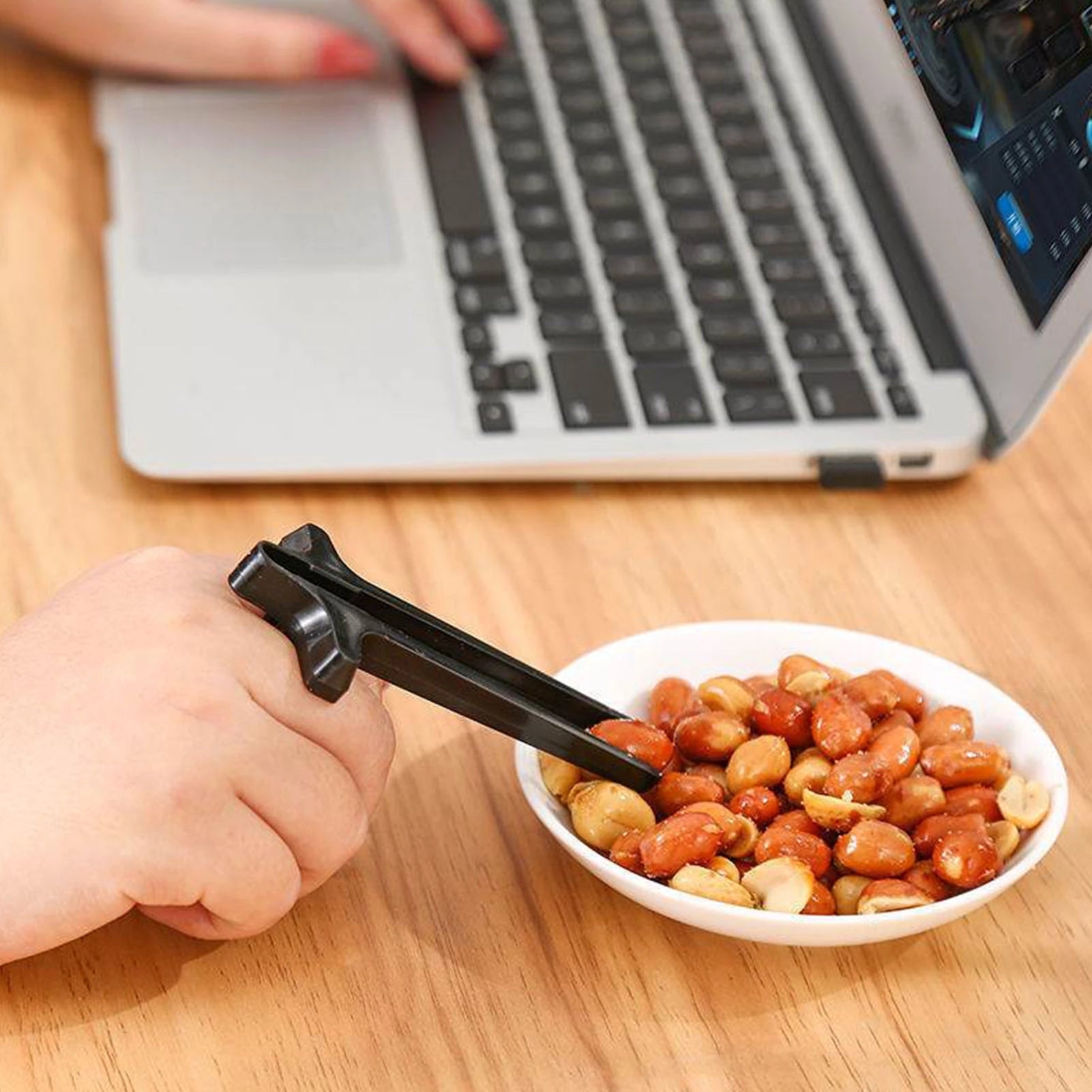 Person using tongs to pick up peanuts from a bowl on a desk with a laptop in the background